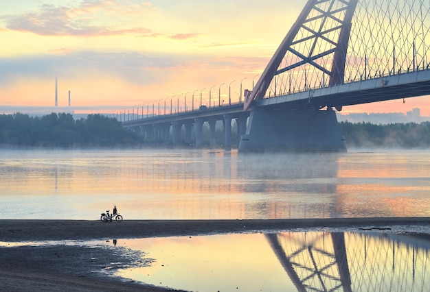 Bici sulla spiaggia. Car bugrinskij ponte sul grande fiume siberiano Ob nella nebbia mattutina