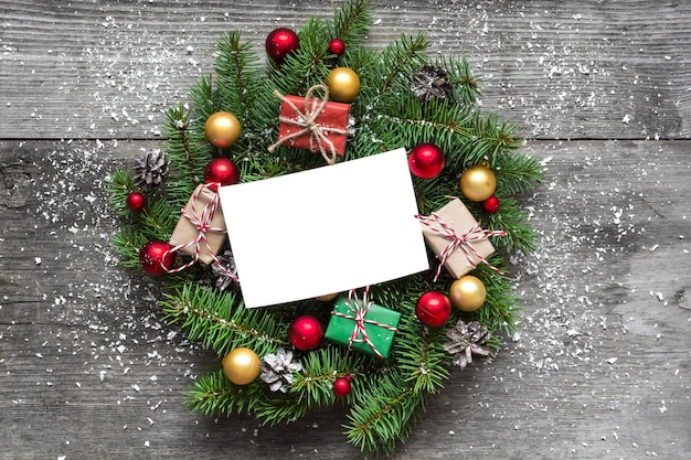Biglietto di auguri vuoto in corona di Natale con decorazioni e scatole regalo su tavola in legno rustico ricoperta di neve. vista dall'alto