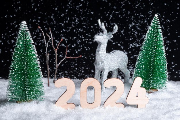 Biglietto di Natale creativo 2024 Un giocattolo splendente cervo d'argento in piedi nella neve nella foresta tra gli alberi di Natale di notte su uno sfondo nero Aiutante di renne di Babbo Natale Felice anno nuovo