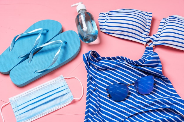 Bikini blu, infradito con maschera protettiva e disinfettante per le mani su sfondo rosa.