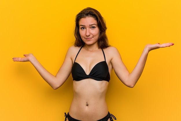 Bikini d'uso della giovane donna europea che dubita e che scrolla le spalle le spalle nel gesto interrogante