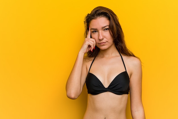 Bikini da portare della giovane donna europea che indica la sua tempia con la barretta, pensante, messa a fuoco su un compito.