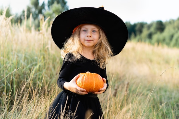 Bimba bionda con un costume nero e un cappello da strega, Halloween con una zucca in mano, un bambino allegro.