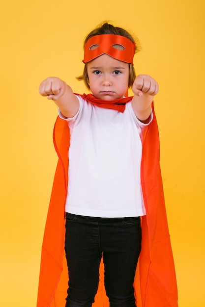 Bimba bionda vestita da supereroina supereroina con mantello rosso e maschera, con le braccia in posizione di volo, su sfondo giallo