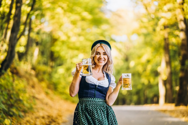 Bionda abbastanza felice in dirndl, abito tradizionale da festa, con due boccali di birra all'aperto nella foresta