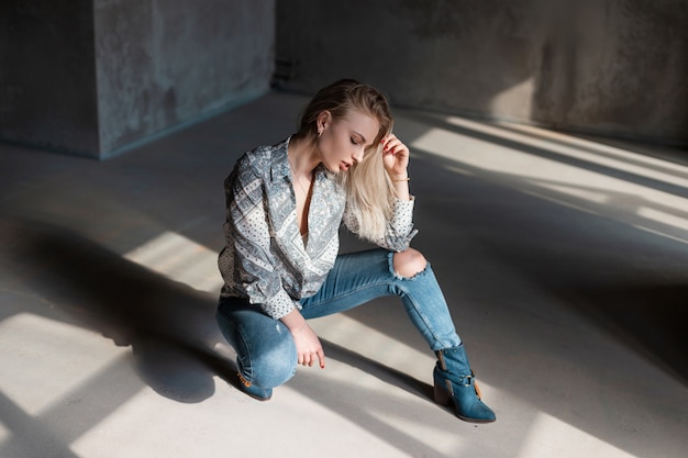 Bionda abbastanza moderna della giovane donna in camicia estiva alla moda in jeans strappati blu in stivali da cowboy alla moda in posa seduta al chiuso con luce solare