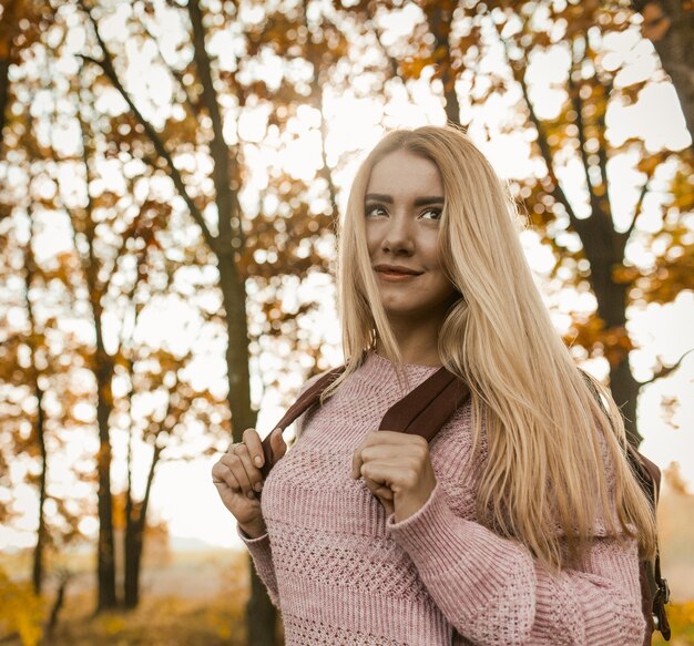 Bionda affascinante che cammina con lo zaino attraverso la foresta di autunno