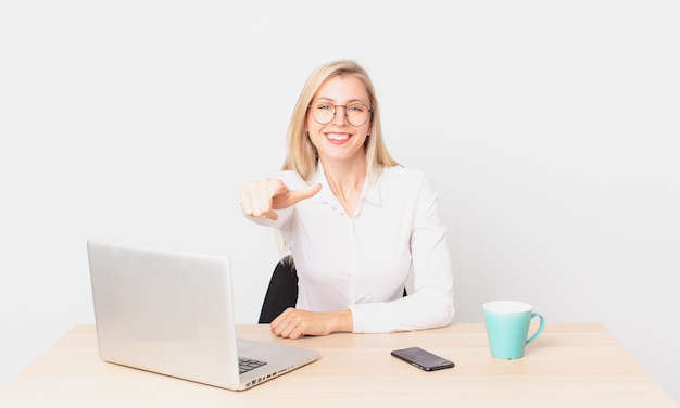 Bionda bella donna giovane donna bionda che punta alla telecamera scegliendo te e lavorando con un laptop