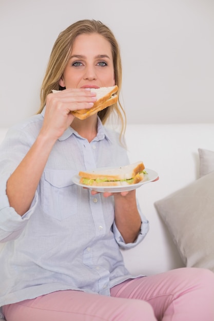 Bionda casuale felice che si siede sul divano che mangia panino