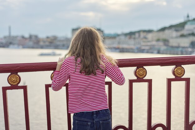 Bionda del bambino con capelli lunghi che stanno indietro sul ponte pedonale sopra il fiume. Ragazza sognante che guarda e si gode il tramonto sulla superficie dell'acqua