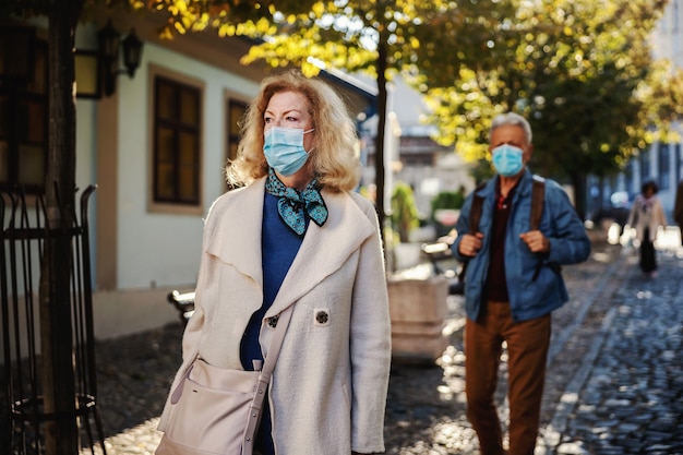 Bionda donna senior con una maschera protettiva sul camminare in centro in una soleggiata giornata autunnale.