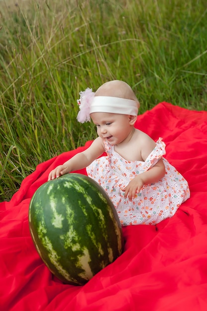 Bionda felice sveglia con la ragazza degli occhi azzurri 8 mesi in erba con l'anguria. Concetto di alimentazione sana, infanzia, maturazione dei frutti, genitorialità, raccolto stagionale. Perfetto bambino caucasico in natura estate