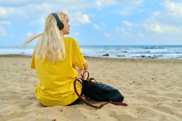 Bionda femminile teenager moderna in cuffie con uno zaino che si siede sulla spiaggia