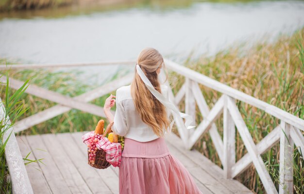 Bionda giovane donna con i capelli lunghi in abiti vintage stile retrò si leva in piedi con un cestino da picnic su un molo di legno del lago