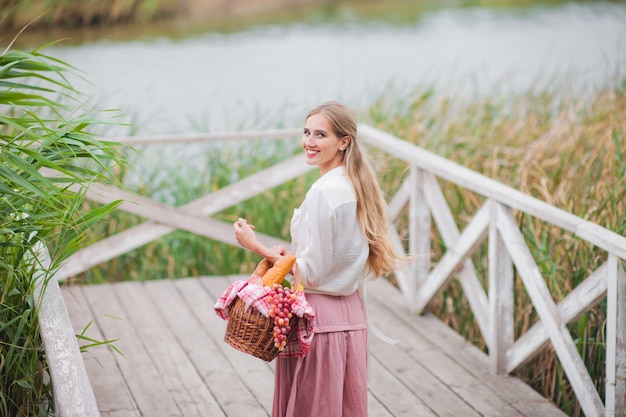 Bionda giovane donna con i capelli lunghi in abiti vintage stile retrò si leva in piedi con un cestino da picnic su un molo di legno del lago