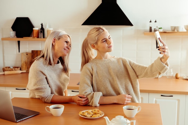 Bionda giovane madre e figlia felici in cucina che fanno selfie per il social network blogger blog vlogger di famiglia