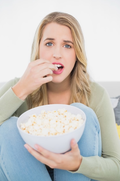 Bionda graziosa sorpresa che mangia popcorn che si siede sul sofà accogliente