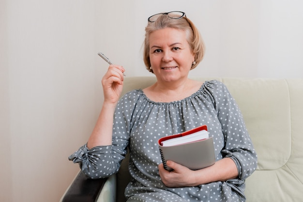 Bionda imprenditrice, sorridente e anziana, con gli occhiali, leggendo e navigando con un'agenda quotidiana e una penna a sfera. Ritratto di un bel pensionato ben curato 50-60 anni in camera.