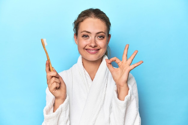 Bionda in accappatoio con uno spazzolino da denti in studio blu allegra e sicura mostrando un gesto ok