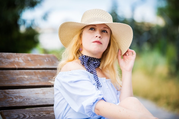 Bionda in un vestito blu e un bel cappello in posa su una panchina in un parco cittadino