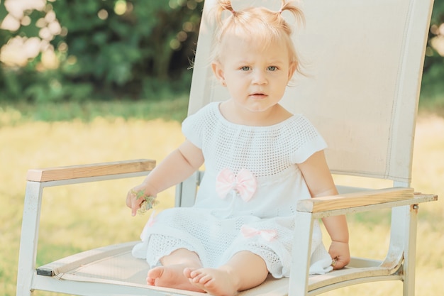 Bionda infantile caucasica della ragazza che si siede su una sedia nel giardino di estate. Foto di alta qualità