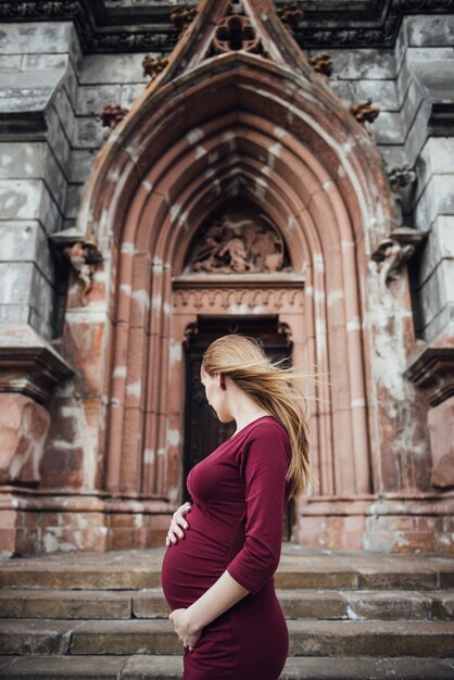 Bionda ragazza incinta in abito bordeaux. Nella grande città. Capelli lunghi. 9 mesi di attesa. La felicità di essere una madre. Sullo sfondo di un bellissimo edificio antico.