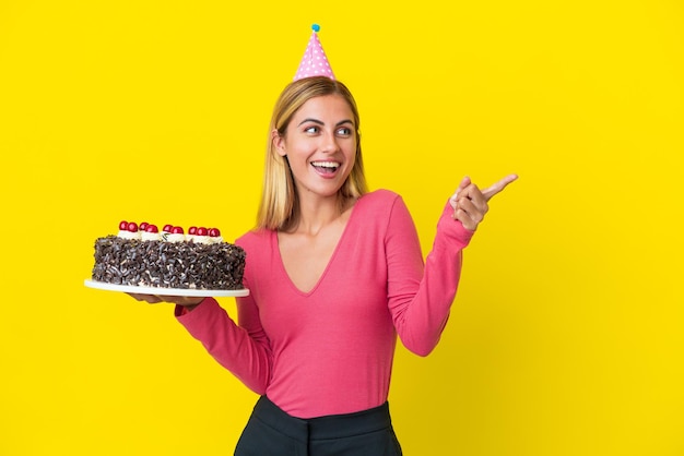 Bionda ragazza uruguaiana con torta di compleanno isolata su sfondo giallo puntando il dito sul lato e presentando un prodotto