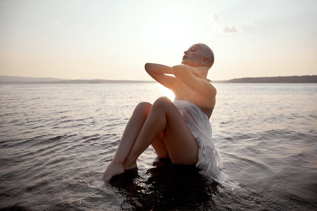 Bionda sexy nuda d'arte con taglio di capelli corto è seduto in acqua sulla spiaggia di riva del lago al tramonto. Capelli bagnati e corpo di donna. Vacanza al mare appartata