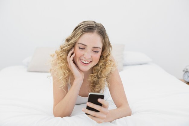 Bionda sorridente graziosa che si trova sul letto facendo uso dello smartphone