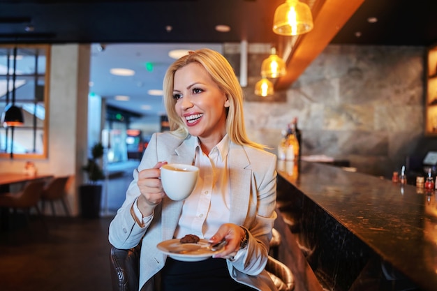 Bionda sorridente in abiti da cerimonia seduto in un bar di un hotel di lusso, bevendo caffè e aspettando il check-in.