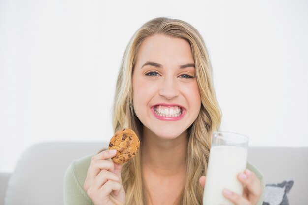 Bionda sveglia felice che mangia biscotto con latte che si siede sul sofà accogliente