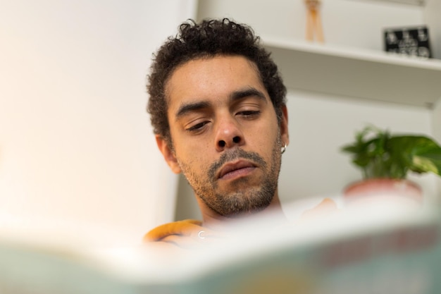 Biracial uomo che legge un libro seduto sul divano di casa in abbigliamento casual Primo piano