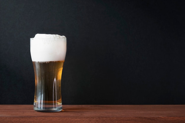 Birra fredda schiumosa in boccale di birra di vetro sulla tavola di legno in un pub. Muro nero scuro come sfondo per testo, menu, mockup