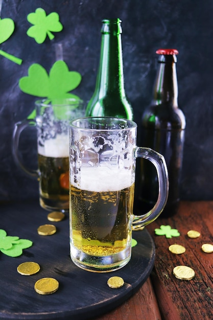 Birra schiumosa di giorno di Patrick in tazze di vetro e una bottiglia di monete d'oro su un trifoglio verde della tavola di legno