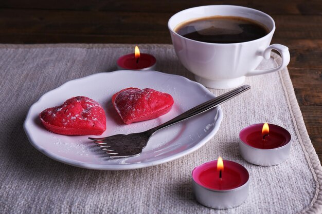 Biscotti a forma di cuore nel piatto con una tazza di caffè e candele sul tovagliolo, su fondo rustico di assi di legno