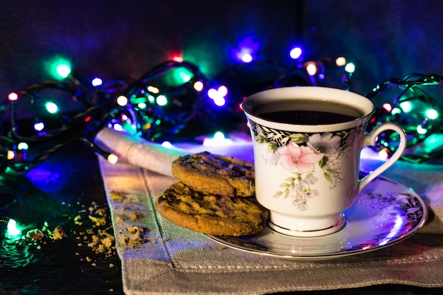 Biscotti al cioccolato e una tazza di tè su sfondo scuro con luci colorate.