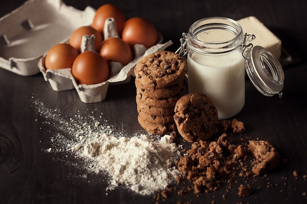 Biscotti al cioccolato su tavola in legno rustico con bottiglia di latte, farina bianca, uova fresche, burro e briciole. Dolce sano.