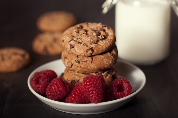 Biscotti al cioccolato sul piatto bianco scuro vecchio tavolo in legno con lampone rosso e latte. Colazione deliziosa.