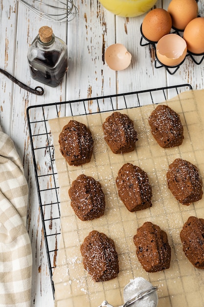 Biscotti casalinghi di Madeleine del cioccolato del forno sullo scaffale di raffreddamento con gli ingredienti e gli utensili su di legno rustico