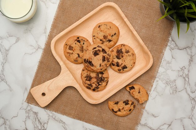 Biscotti con gocce di cioccolato appena sfornati su un piatto di legno una tazza di latte e una pianta decorativa su un tavolo di marmo