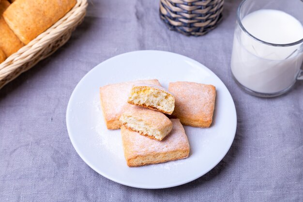 Biscotti con zucchero a velo su un piatto bianco. Sullo sfondo un cesto di biscotti e una tazza di latte. Avvicinamento.