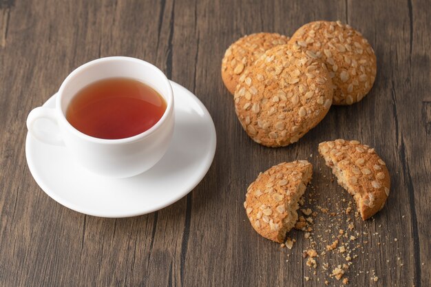 Biscotti di farina d'avena con semi vicino a una tazza bianca di tè nero su una superficie di legno