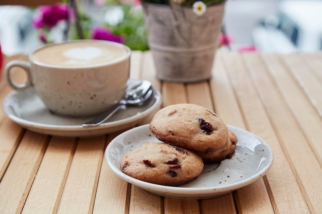 Biscotti di mirtilli rossi fatti in casa naturali senza zucchero sul piatto e cappuccino in tazza di ceramica bianca sul tavolo di legno sulla terrazza esterna Colazione estetica nella caffetteria