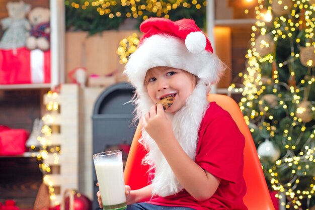 Biscotti e latte di Natale. Piccolo bambino di Babbo Natale con barba e baffi. Babbo Natale in casa. Santa