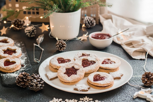 Biscotti pronti Linzer con marmellata di frutti di bosco su un piatto e decorazioni natalizie su uno sfondo scuro. Cucinare dolcetti natalizi. Stile di vita