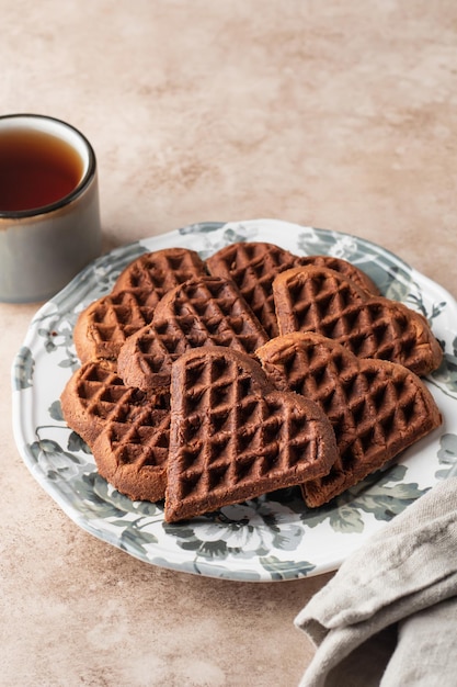 Biscotti waffle al cioccolato a forma di cuore in piatto festivo per San Valentino con tè e tovagliolo su sfondo beige Dolci dolci con amore Regalo per San Valentino