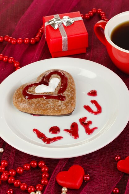Biscotto al cioccolato a forma di cuore con una tazza di caffè sul primo piano rosa della tovaglia