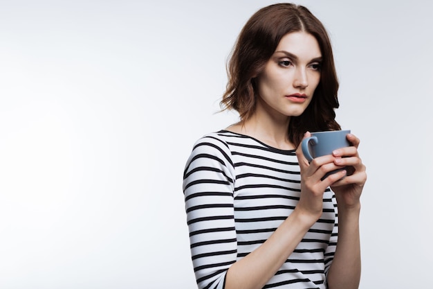 Bisogno di dormire di più. Piacevole donna dai capelli ramati in un pullover a righe che beve caffè, con l'aria esausta dopo una notte insonne