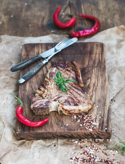 Bistecca di bistecca con l'osso cotta sulla tavola di servizio con peperoncino rosso, spezie e rosmarino fresco su carta oleosa artigianale, in legno rustico