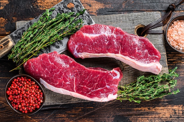 Bistecca di carne di manzo controfiletto crudo su un tagliere di legno con erbe aromatiche. Fondo in legno scuro. Vista dall'alto.